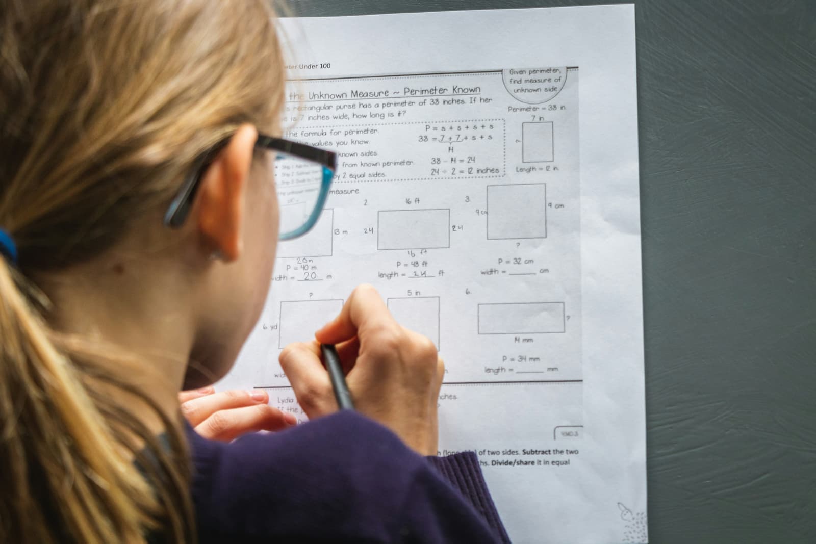 An over-the-shoulder view of a learner in glasses working through perimeter problems on a printed maths worksheet
